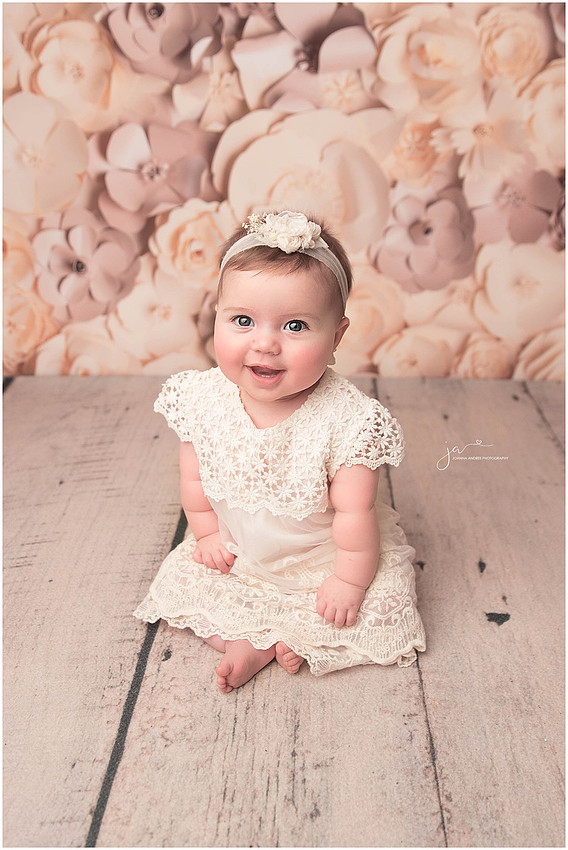 Baby Girl sitting up in a cream dress with flowered backdrop